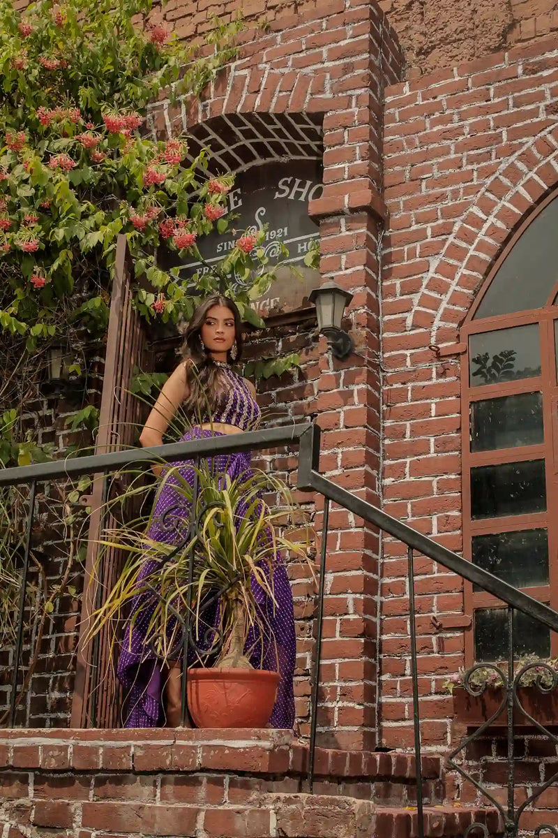 Far-shot portrait of a model on an outdoor balcony wearing a deep purple ethnic co-ord set with traditional Indian hand-embroidery.