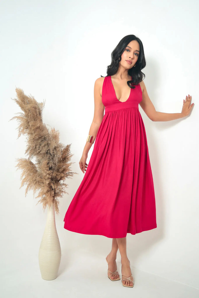 A woman leaning against a white wall styling a vibrant crimson pink twill midi dress with a plunge neckline, gathered empire waist, and flowy silhouette, accessorized with a gold cuff bracelet.