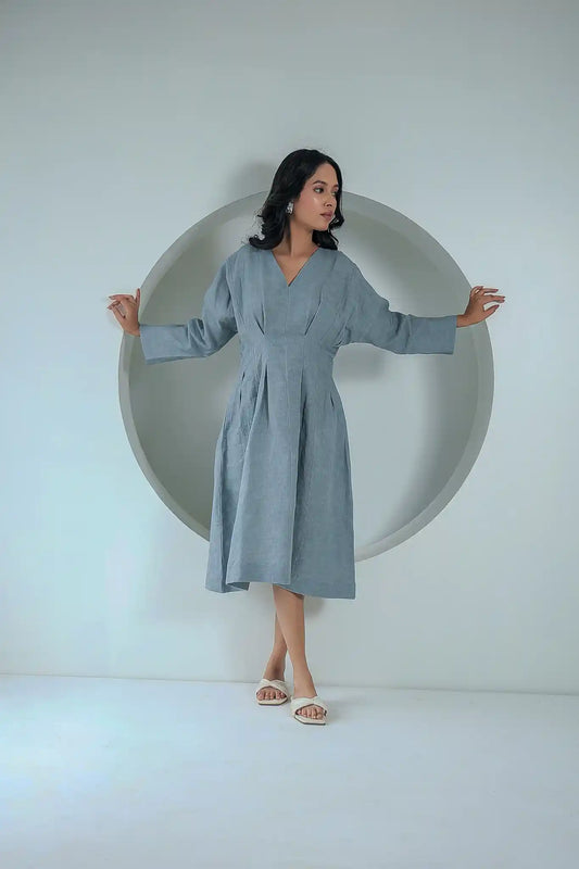 Full-length front view of a woman wearing a grey box-pleated linen midi dress with long sleeves and a V-neckline, standing against a white circular studio backdrop.