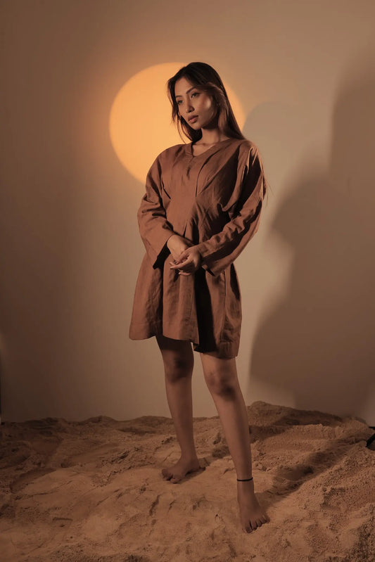 Barefoot model standing on sand under a circular warm spotlight, showcasing the full loose silhouette and front box pleat detail of the nutshell brown short linen dress.