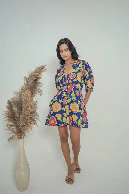 Full-length view of a model wearing the Tea & Dove Celestial Bloom short linen dress in galactic cobalt blue with bright floral prints, featuring a V-neckline and side pockets.