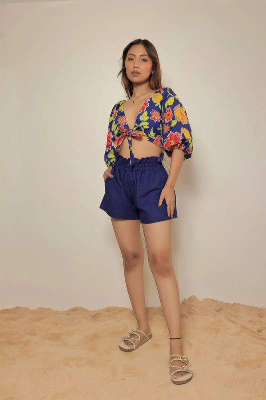 Full-length front view of a model standing on sand wearing the Tea & Dove Cloud Knot cobalt blue linen shorts and matching floral puff-sleeve front-knot crop top set.