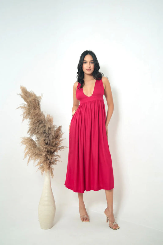 Full-length studio portrait of a woman posing in the Tea & Dove Elysian Gown in crimson pink twill, featuring a deep V-neckline, empire waist, and a flowing midi-length skirt next to a white vase with pampas grass.
