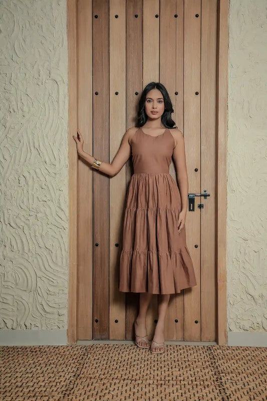 A woman posing in front of a rustic wooden door wearing a sleeveless, hazelnut-brown tiered midi dress with a fitted bodice and round neckline, paired with a gold cuff bracelet and samish light brown colour low heels.