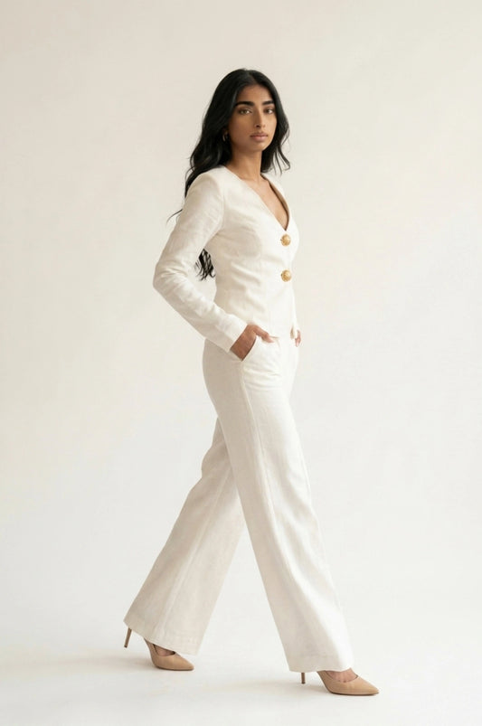 Full-length side profile view of a woman posing in motion against a white background. She is wearing a fitted ivory suit jacket featuring gold buttons, wide-leg trousers with a crease down the front, and nude high heels, with one hand tucked into her pocket.