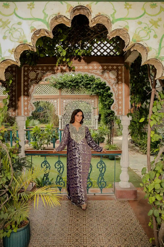 tea-&-dove-sitara-purple-&-gold-lurex-posed-front-view.webp: Full-length shot of a model wearing a luxurious deep purple and gold shimmering lurex kaftan under an ornate garden archway.