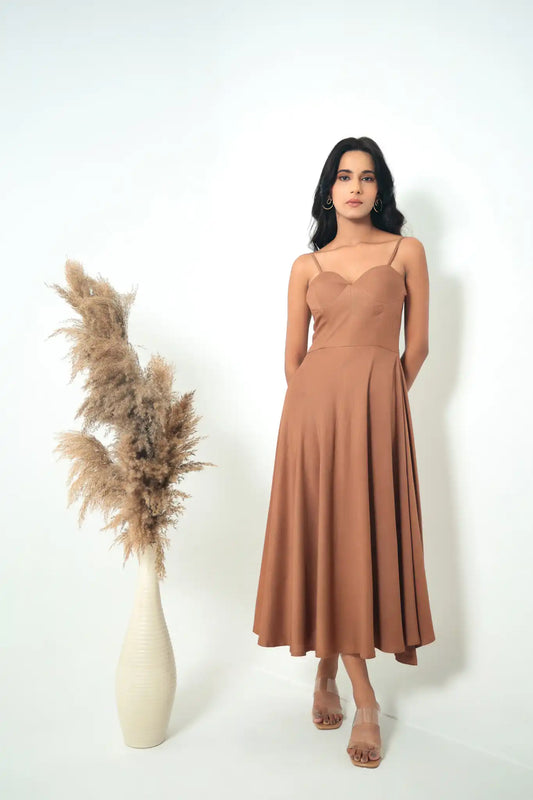 Full-length studio shot of a model wearing the tea & dove Twilight Embrace midi dress in nutshell brown, featuring a sweetheart neckline and A-line silhouette, styled next to a tall vase with dried pampas grass.