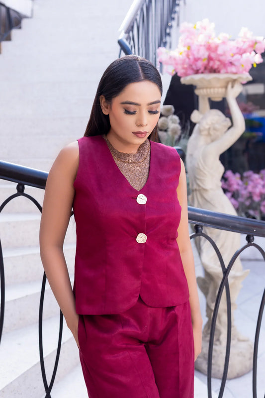 Medium front shot of a woman looking down while leaning against a black railing outdoors. She wears a tailored burgundy suede vest over a shimmering bronze mesh top, paired with matching burgundy suede trousers.